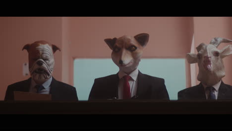 A girl sits at a table and strange figures in animal masks observes her from behind the glass