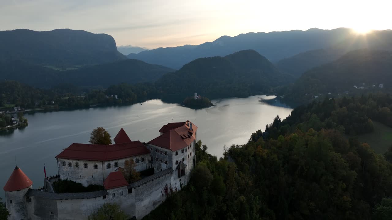 Un'ampia ripresa aerea in allontanamento rivela il Lago di Bled e le montagne circostanti con il castello sulla scogliera all'alba