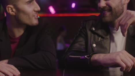 Close-Up-Of-Two-Male-Friends-Toasting-With-Beer-Bottle-And-Talking-Together-While-Sitting-At-Bar-Counter