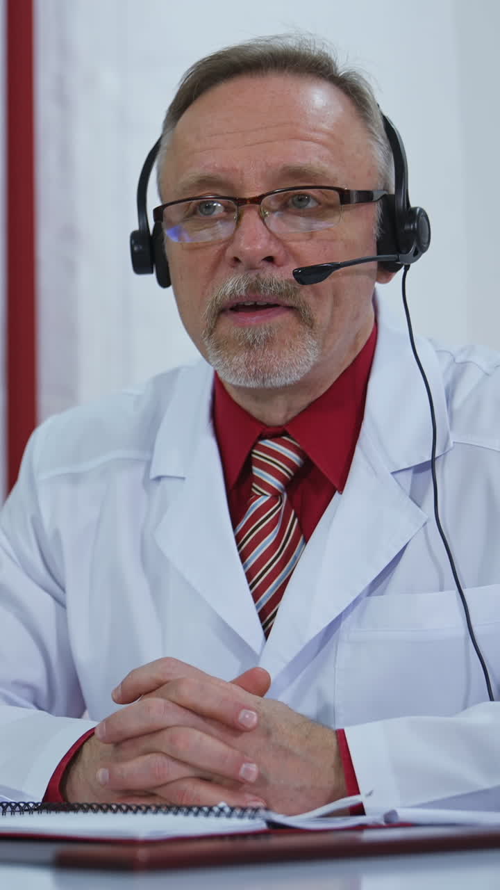 Doctor with headset talking on camera in medical office. Professional doctor in white medical coat sitting at desk and giving online consultation during quarantine. Vertical video