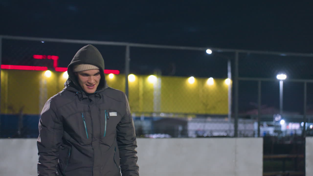 joven con capucha sonriendo con expresión enfocada durante la actividad nocturna en el campo urbano al aire libre, el fondo presenta una valla iluminada, luces amarillas y rojas vibrantes
