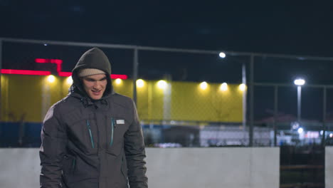 young man in hoodie smiling with focused expression during nighttime activity on outdoor urban field, background features illuminated fence, vibrant yellow and red lights