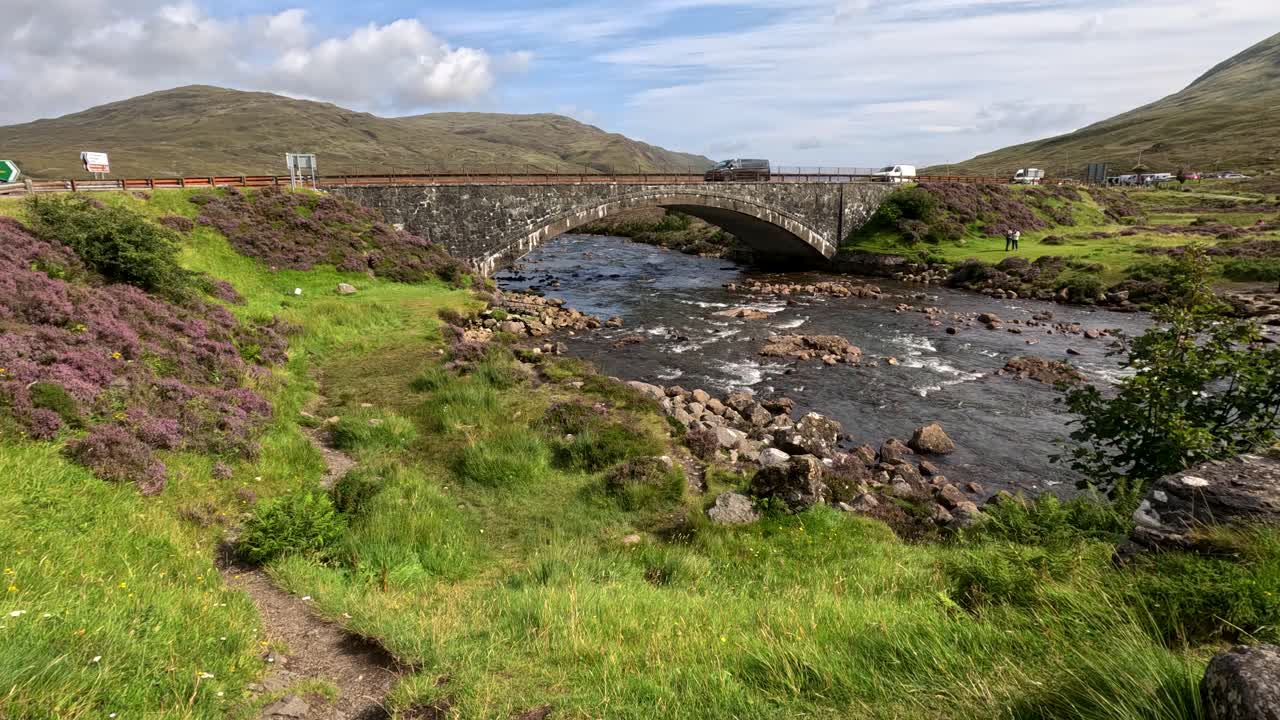 Camera moves slowly along a grassy riverside path toward a stone bridge over a clear mountain stream, with vibrant wildflowers and rolling hills under daylight