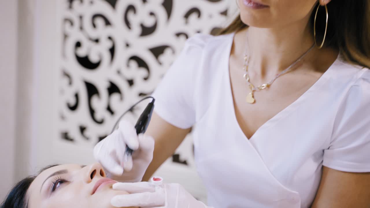 Beautiful cosmetologist applying permanent make-up on lips in a beauty salon. Close-up attractive beautician doing tattooing lips for a female patient.