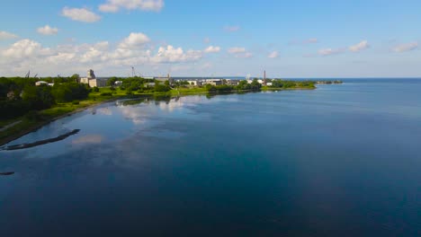 aerial view orbit over kopli peninsula blue mirror like sea water reflecting clouds. coastal maritime industrial area with cranes, warehouses and seaside buildings along green trees, sunny summer day