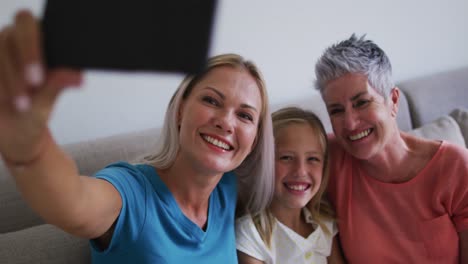 caucasian grandmother, mother and daughter smiling while taking a selfie with smartphone at home