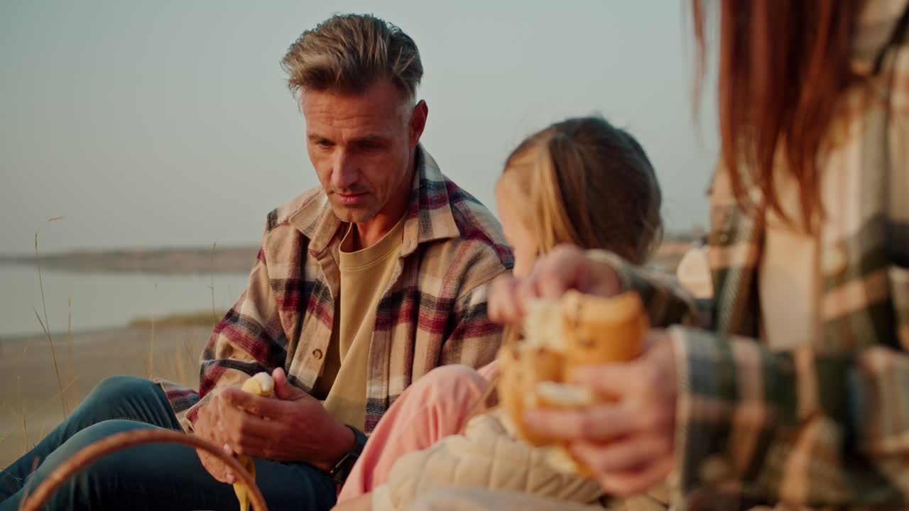 A middle-aged brunette man in a checkered shirt peels a banana fruit for his little daughter during a picnic outside the city in summer. Happy man feeding his little daughter with his wife during a picnic outside the city in summer
