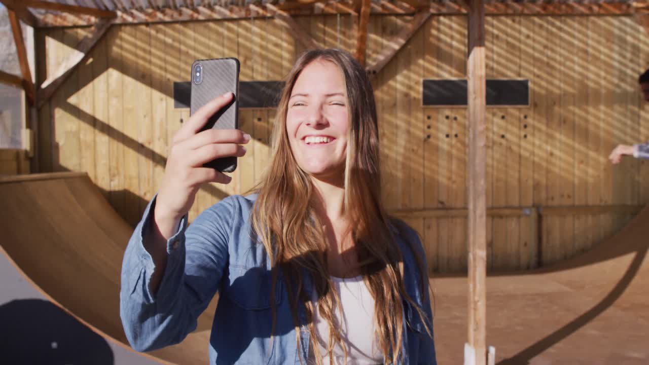 Video iloisesta valkoihoisesta naisrullalautailijasta ottamassa selfietä skeittipuistossa.