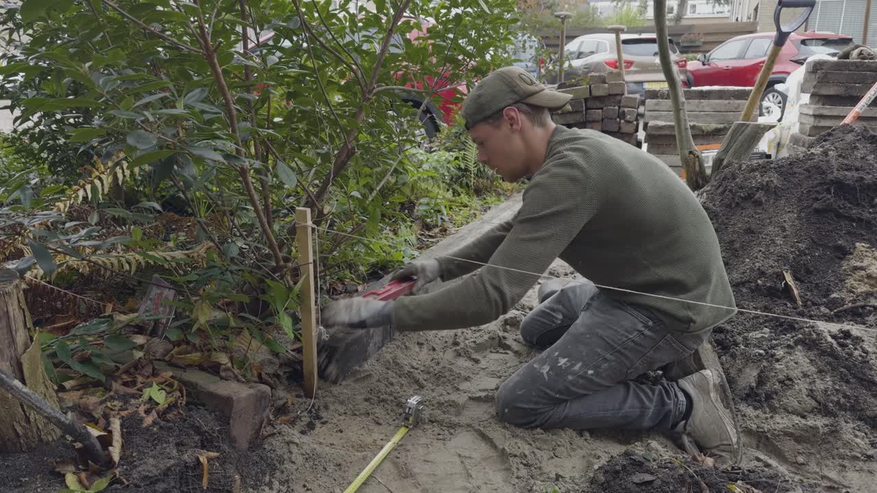 Close-up of a gardener kneeling in the dirt while measuring and placing wooden posts during a garden landscaping job, surrounded by tools, bushes, and soil