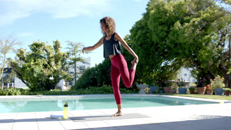 feliz mujer biracial practicando yoga en la piscina, sosteniendo el pie en el jardín soleado, copia espacio, cámara lenta