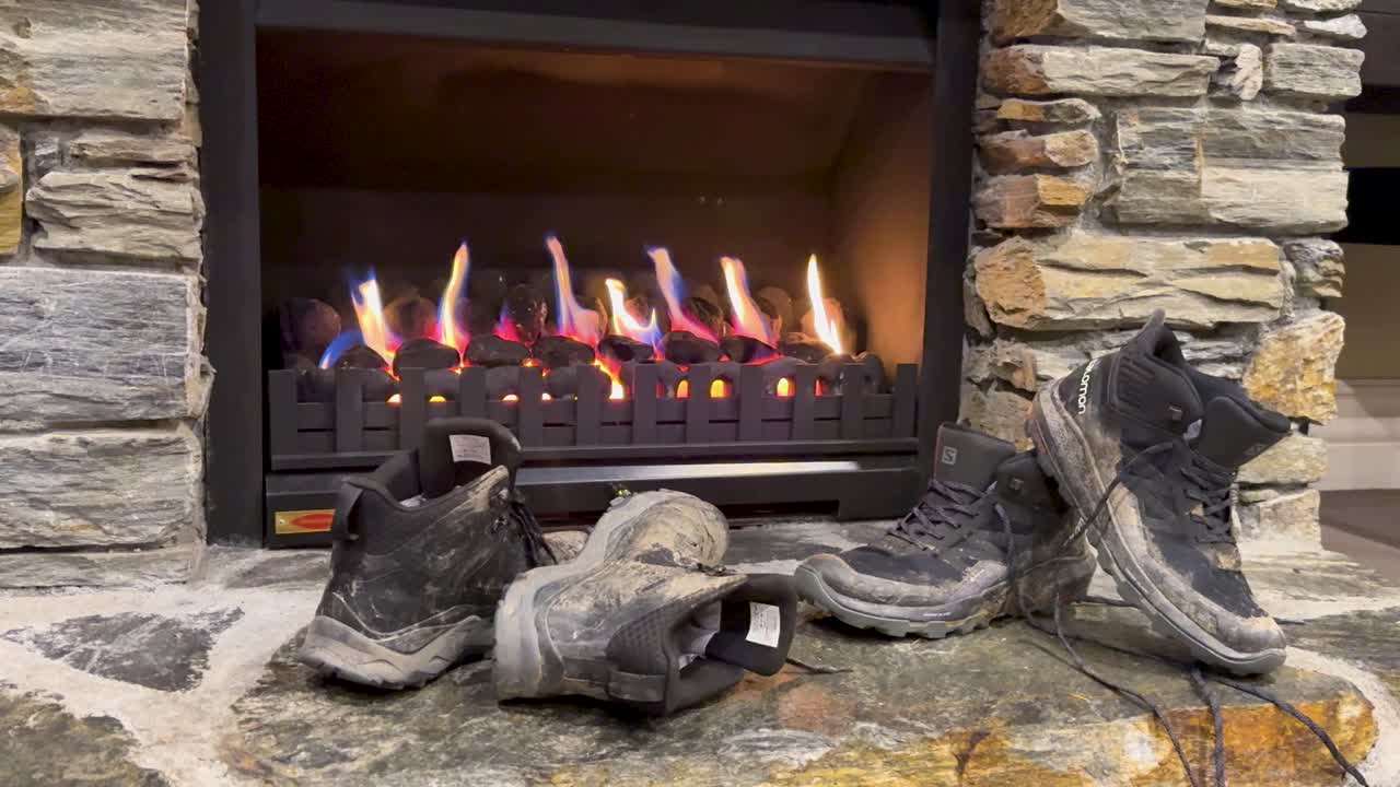 Worn hiking boots dry by a warm fireplace in a rustic stone interior, steady camera
