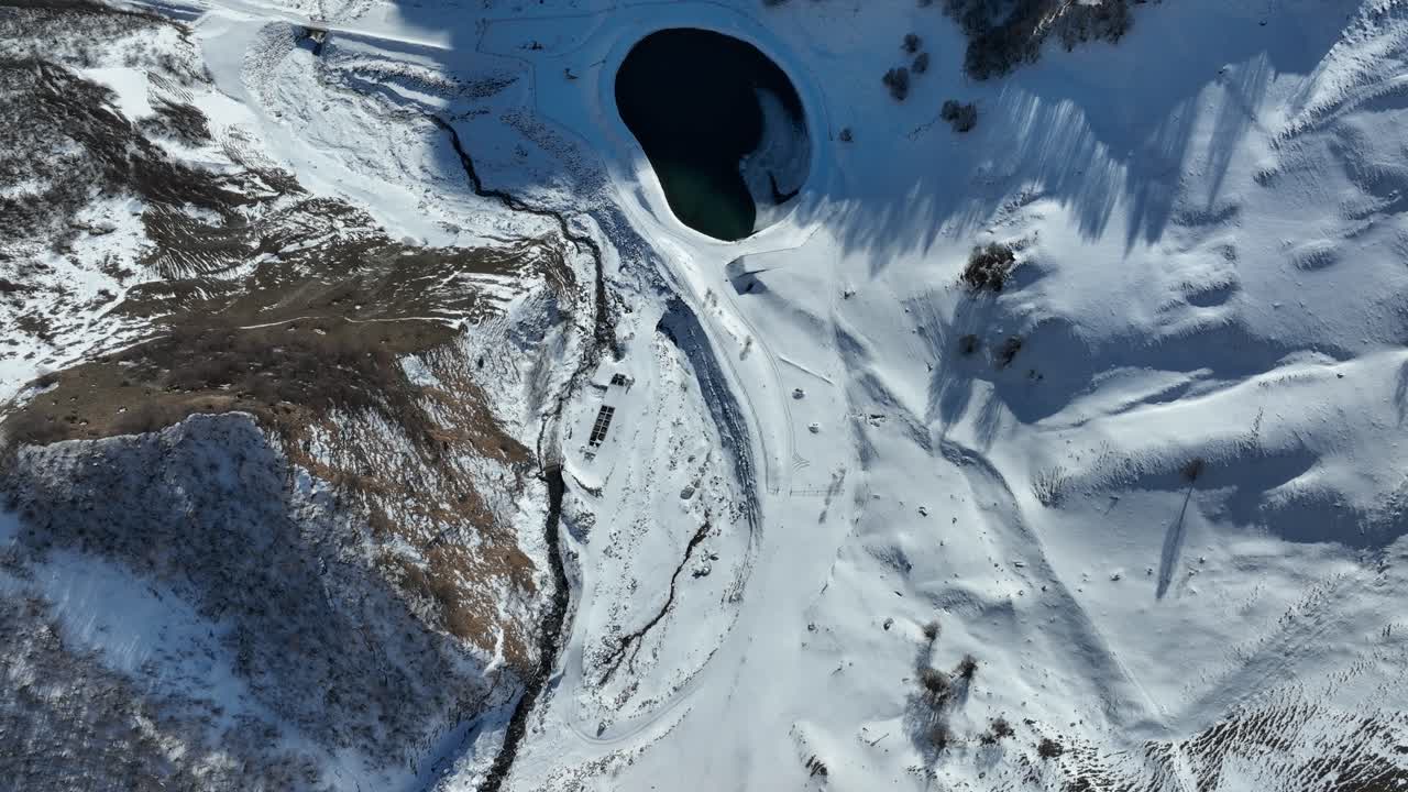 A mesmerizing aerial view of a dark crater lake nestled amidst a snowy mountain range. The contrast between the deep blue water and the surrounding snow-covered peaks creates a breathtaking visual