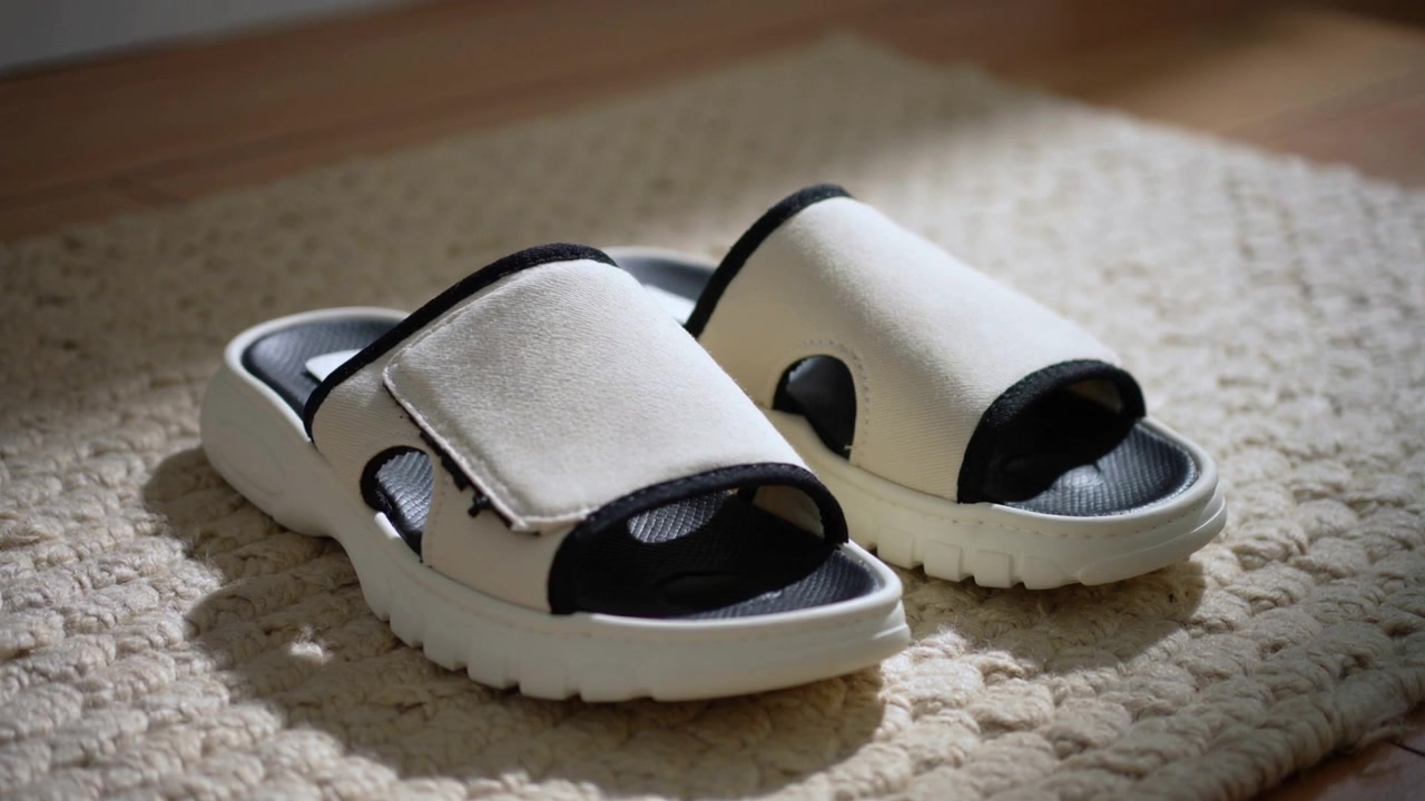 Klipp som viser et par hvite sandaler som hviler på et teksturert beige teppe ved siden av et tregulv.