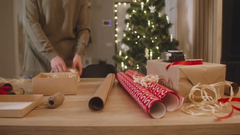 Die-Kamera-Konzentriert-Sich-Auf-Die-Hand-Der-Frau,-Die-Weihnachtsgeschenke-Auf-Einem-Tisch-In-Einem-Mit-Einem-Weihnachtsbaum-Geschmückten-Raum-Einwickelt