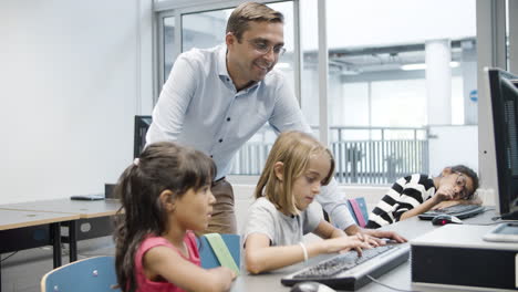 Niñas-Multiétnicas-Escribiendo-En-El-Teclado-Mientras-Un-Profesor-Masculino-Las-Observa