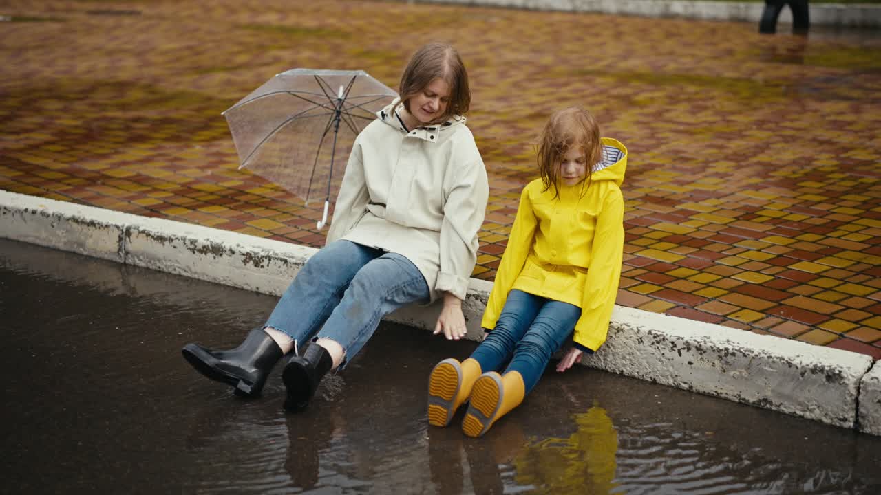 A happy blonde woman in a white jacket and her teenage daughter are sitting on the curb and putting their feet in rubber boots in a puddle after the rain while walking