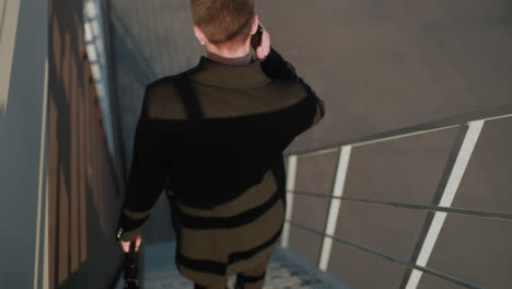 back view man black coat descending staircase on phone call holding briefcase, conversing, modern architecture in background , as strip reflections is seen on his cloth