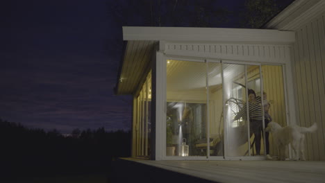 mother opening sliding door at night to let a dog inside while family is gathered indoors