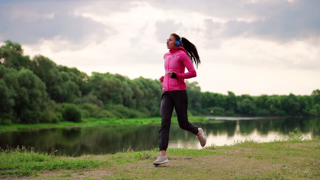 A girl in a pink jacket and black pants runs near the river in headphones preparing for the marathon