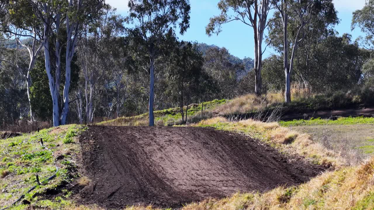 Motorbike rider jumps dirt track berm, bright daylight, rural landscape, static wide shot