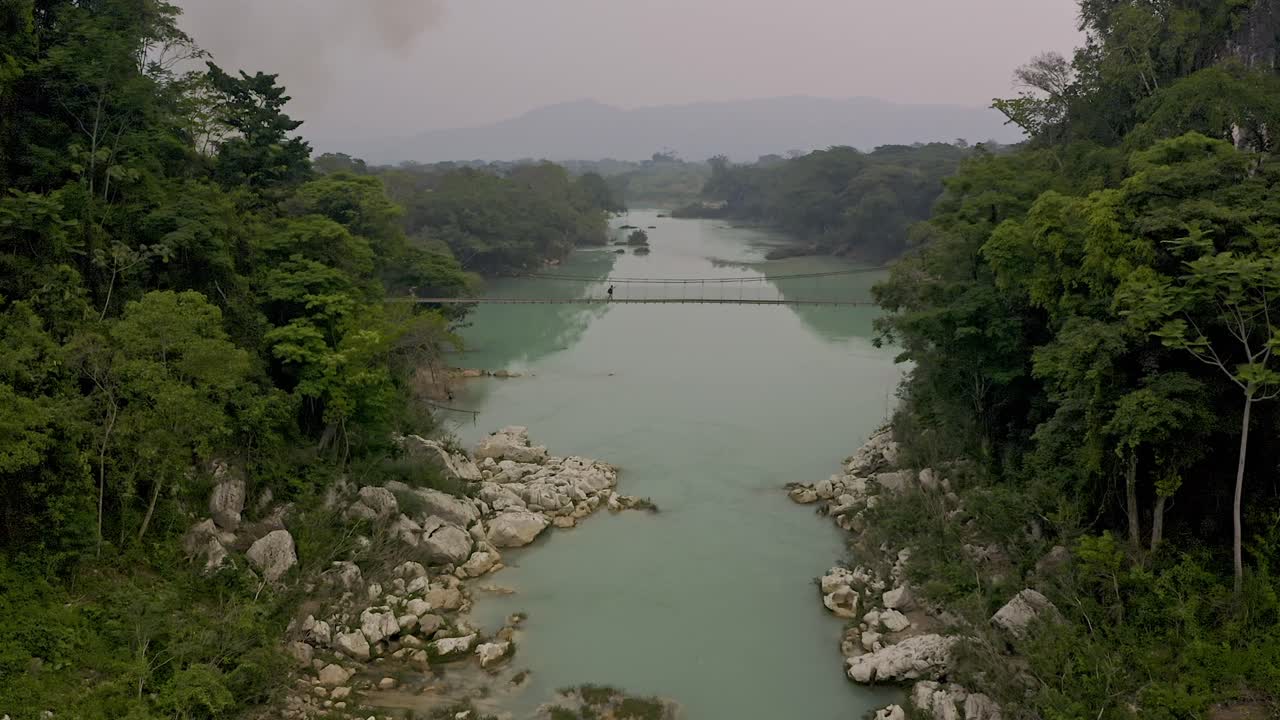 Aerial Drone shot of the Agua Clara, in Chiapas, México. The light-colored river water is spanned by a long, rustic footbridge, on which a lone man is seen crossing It