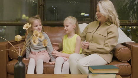 happy grandmother sitting on sofa and hugging her two little granddaughters at home 1