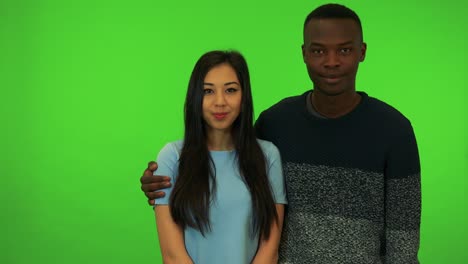 a young black man wraps his arm around a young asian woman's shoulders, they smile at the camera - green screen studio