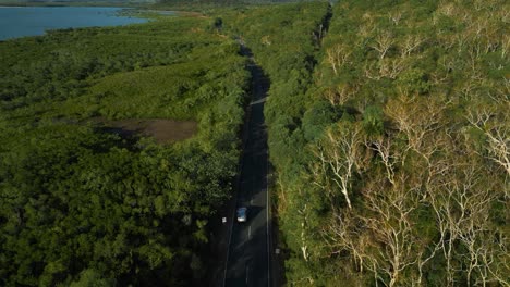 Modern-silver-car-driving-on-road-along-the-seaside-coast-with-sandy-beach-and-lush-green-tree-forest-and-blue-sea-waves