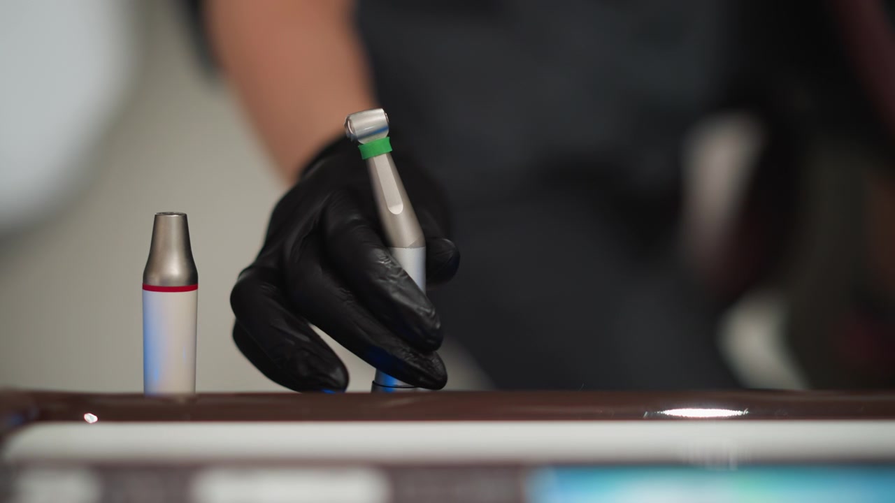 Primo piano di mano guantata che afferra strumenti dentali sul bancone. Una persona che indossa guanti neri e abiti scuri interagisce con due strumenti metallici con dettagli rossi in un ambiente interno.