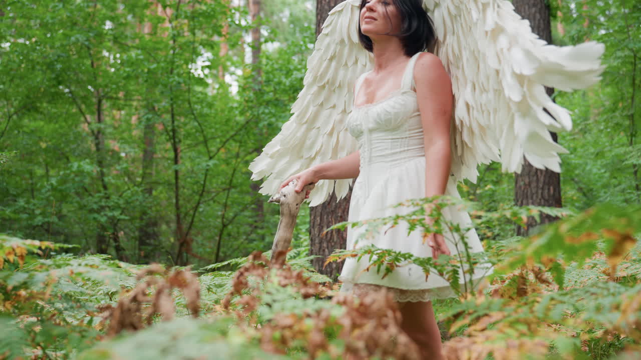 Actress dressed in white angel costume walks gracefully through lush woodland, gently touching green plants and ferns, surrounded by tall trees, creating magical peaceful scene filled with beauty