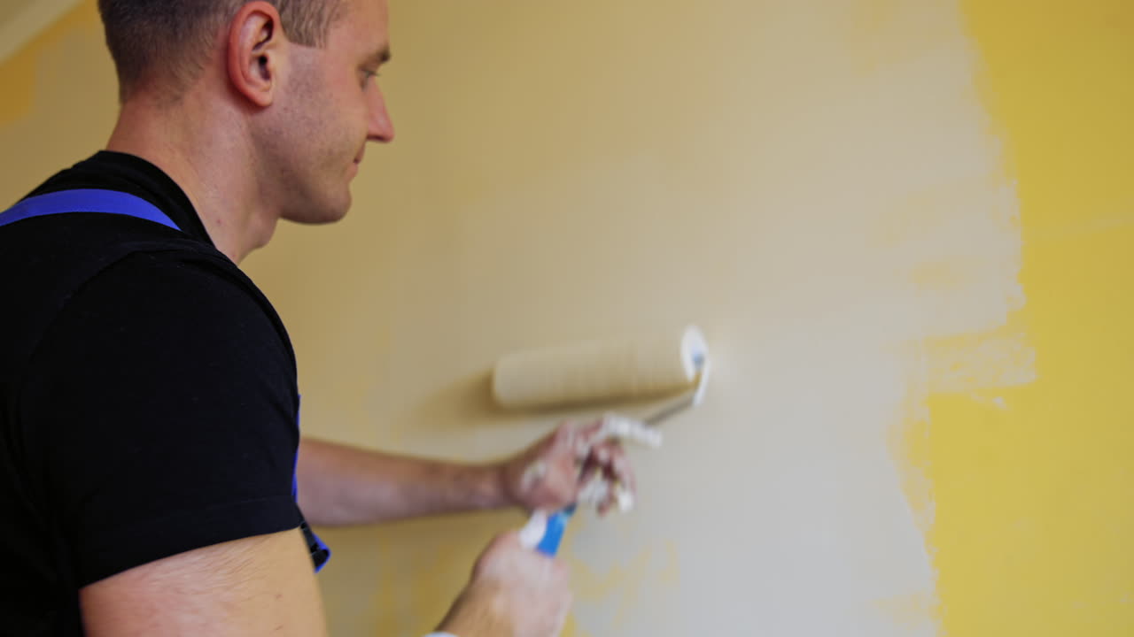 Professional young worker painting walls indoors. Paint roller in man's hands. Craftsman doing makeover in the room. Painting services.