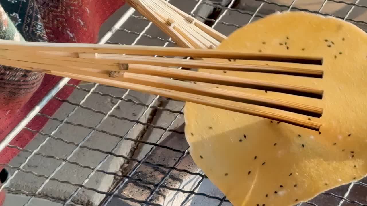 Close-up of a rice cracker being flipped on a grill using bamboo tongs over open flames.