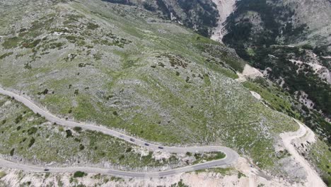 Drone-view-in-Albania-flying-over-a-green-and-rocky-mountain-with-serpent-road-climbing-a-port-with-cars-driving-down-on-a-cloudy-day