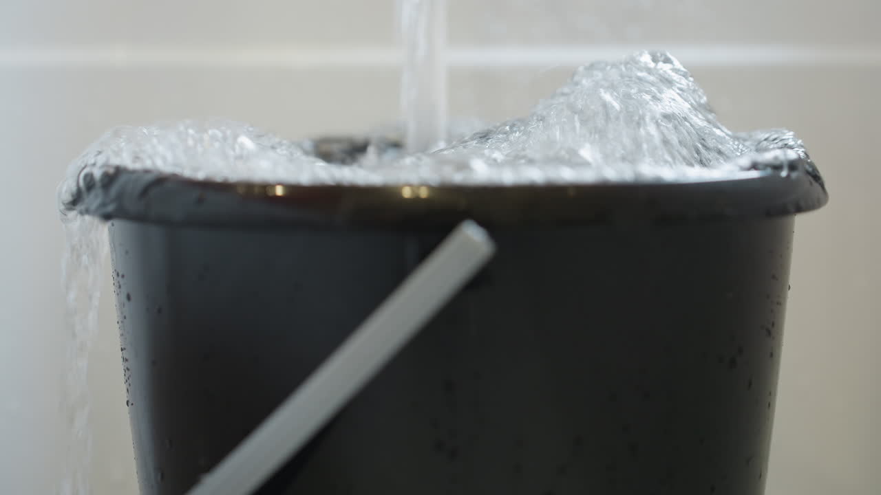 Close up of black bucket filled with water as stream pours inside, liquid overflowing edges with bubbles forming on surface, concept of waste, cleaning, household use and water management