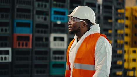 a man with black skin in a white protective uniform in an orange vest in transparent safety glasses walks along yellow and black boxes at a waste processing and sorting plant