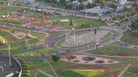 plaza de los mástiles in posadas features tall flagpoles, statues, and a traffic circle, surrounded by roads, parks, trees, and sport courts, drone pull out, wide urban perspective