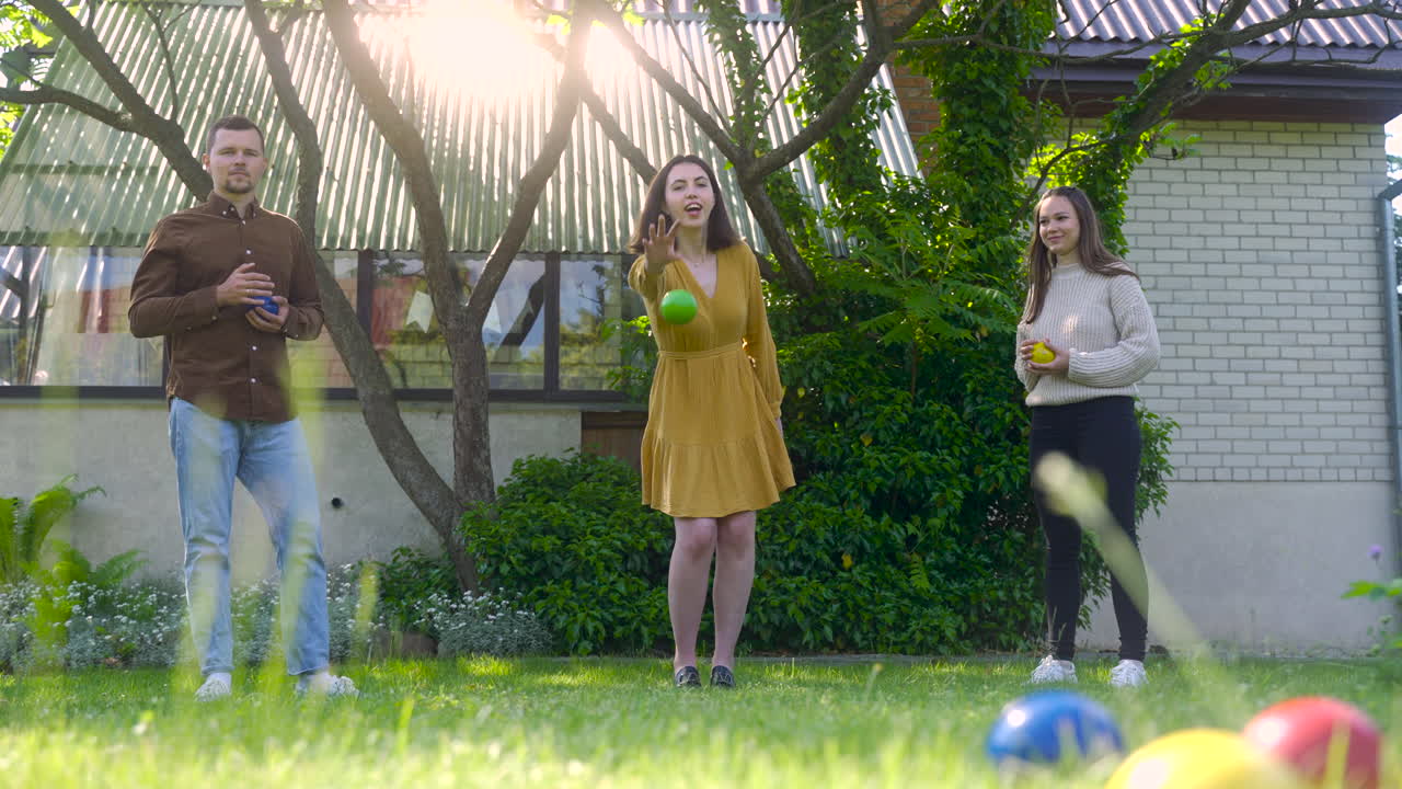 vista frontal de una joven caucásica lanzando una pelota de petanca verde en el parque en un día soleado mientras sus amigos esperan su turno