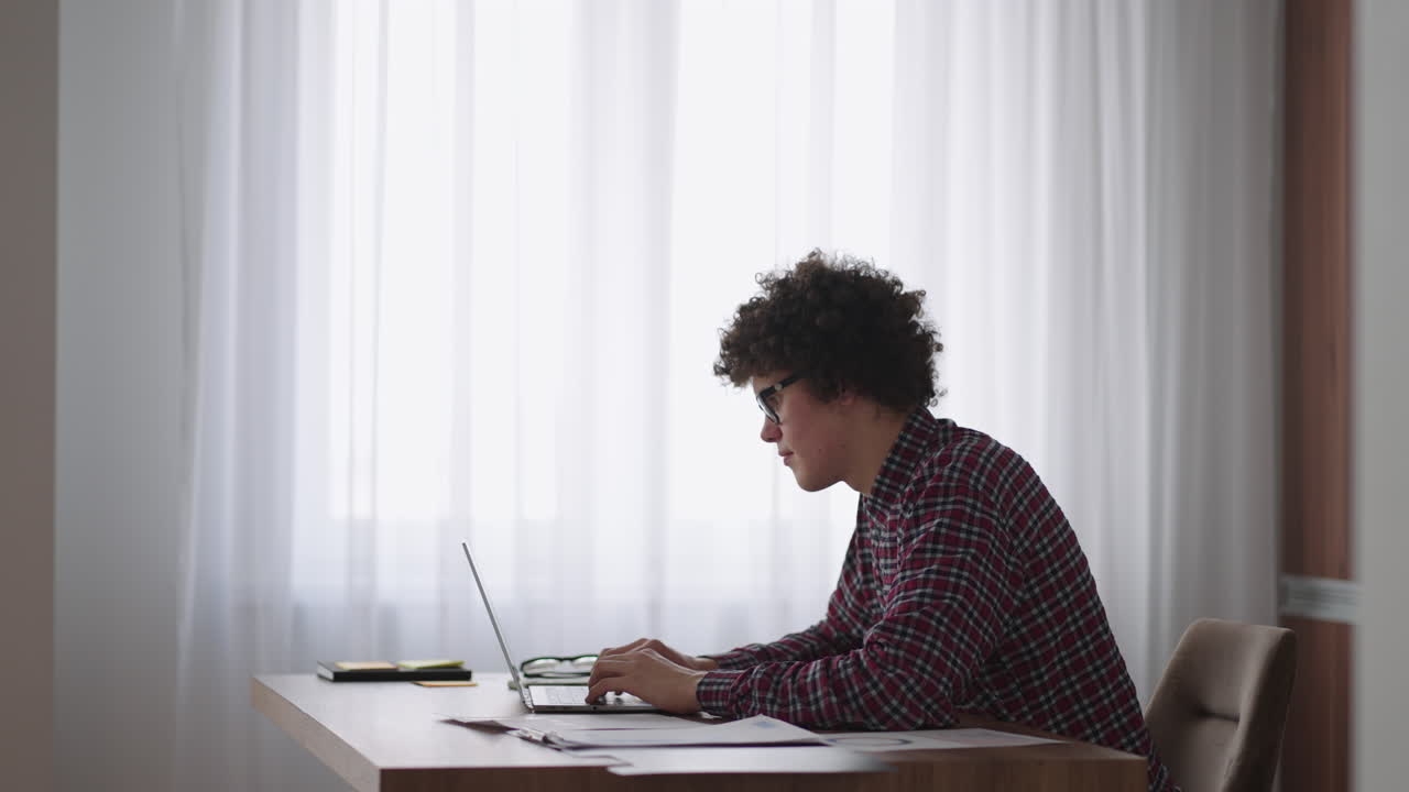 A Curly man with a serious look works at a laptop sitting in a modern kitchen. Young man freelancer student using laptop studying online working from home in internet