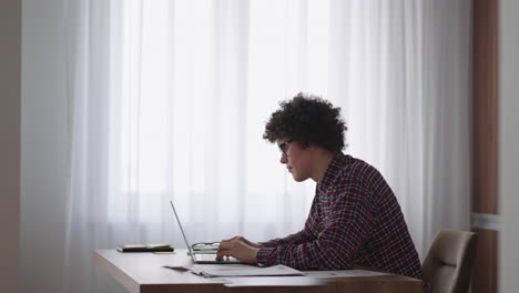 A-Curly-man-with-a-serious-look-works-at-a-laptop-sitting-in-a-modern-kitchen.-Young-man-freelancer-student-using-laptop-studying-online-working-from-home-in-internet