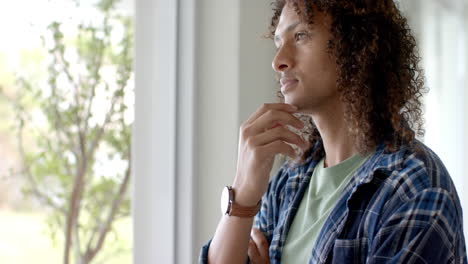 portrait of thoughtful biracial man with long curly hair by window at home, copy space, slow motion