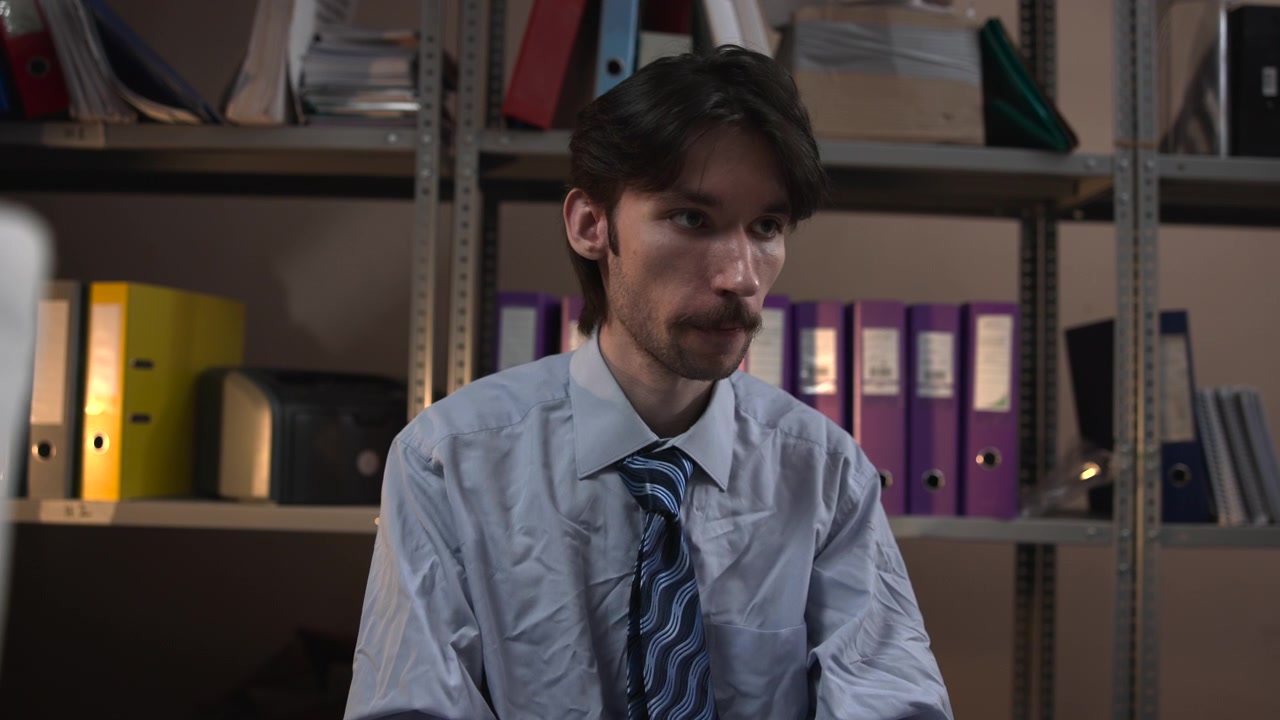 Un joven concentrado se sienta en un escritorio y teclea en un teclado dentro de una sala de archivo con poca luz. Estantes llenos de archivadores y carpetas cubren el fondo.