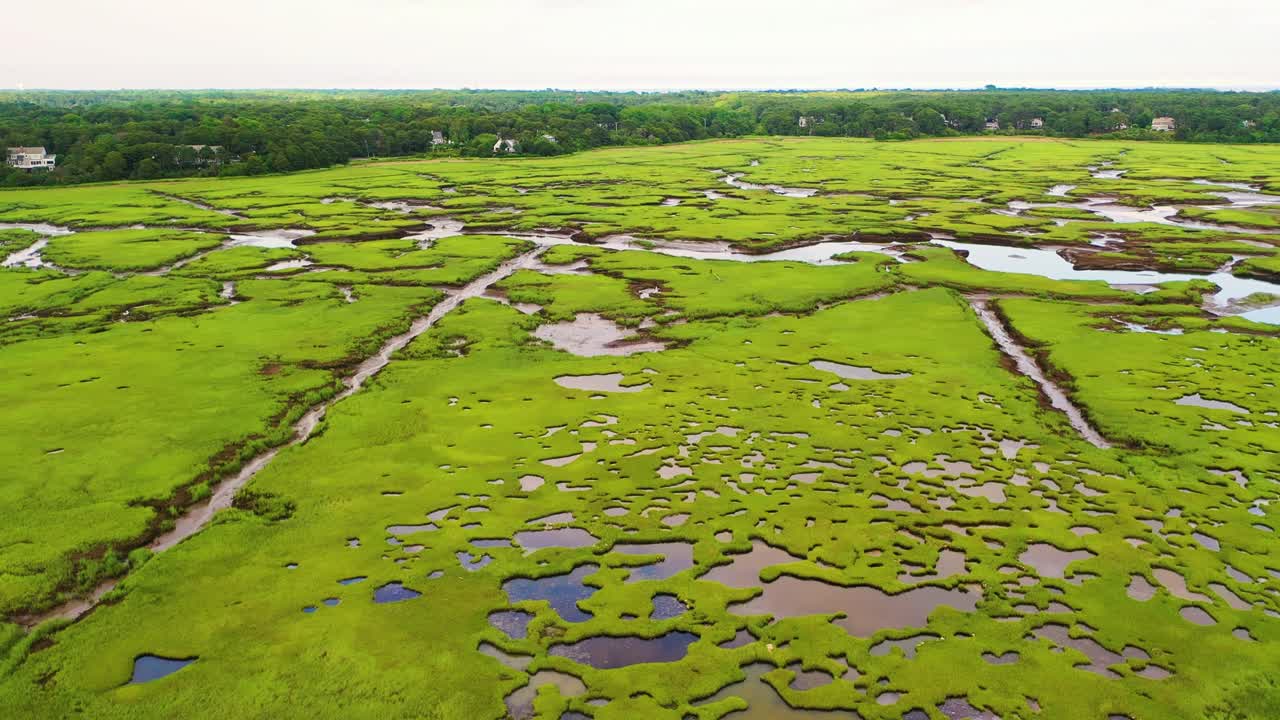 Aerial drone video of Nantucket marsh captures curving tidal creeks, tidal flats, and lush green meadows merging with sandbars and distant tree lines, creating a cinematic coastal landscape