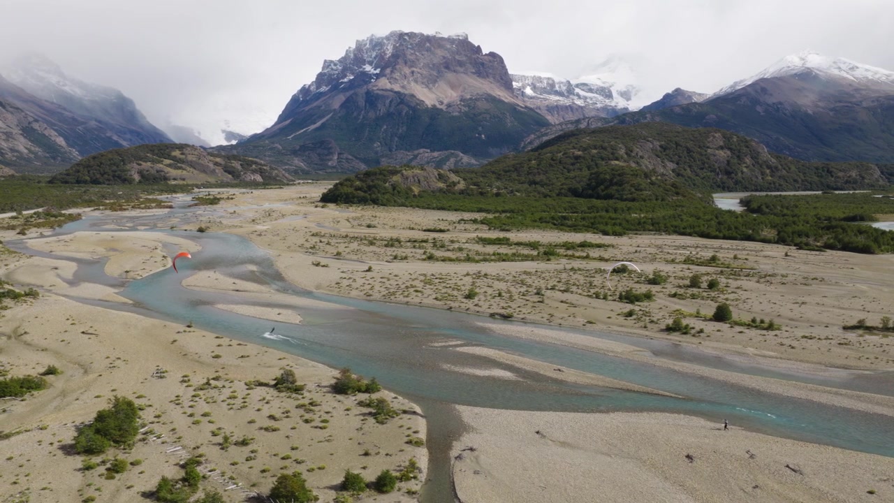 Parapendisti che volano sopra il Río de las Vueltas vicino a El Chaltén, Santa Cruz, Argentina, con canali glaciali intrecciati, ghiaia e il massiccio del Fitz Roy che si erge sopra le Ande meridionali, ripresa aerea con parallasse