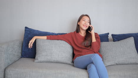 beautiful smiling woman talking on cell phone, smiling asian thai girl sitting on the couch in casual clothes and having mobile phone conversation holding smartphone near ear