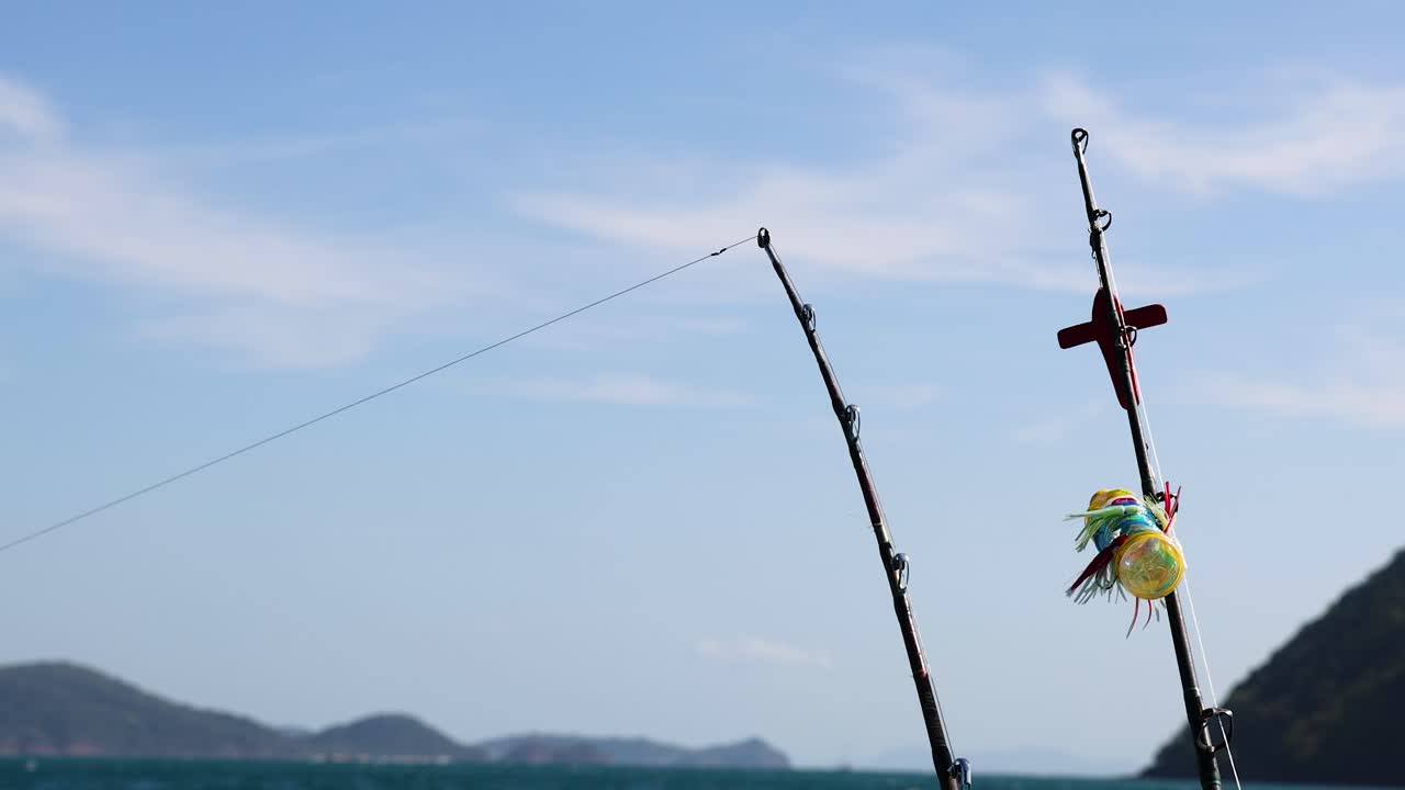 A fishing rod gently sways on a boat against a clear blue sky and distant hills in Phuket, Thailand
