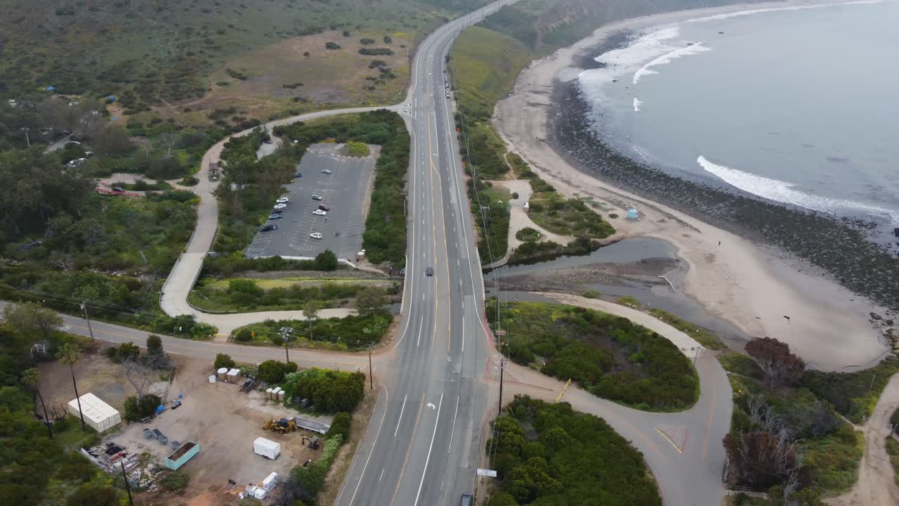 Overhead shot of car driving towards the camera up the Pacific Coast Highway on the California coast. Contrasted by lush greenery and waves crashing onto the beach below. 4K drone road trip shot.