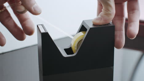 man pulling out transparent duct tape by hand and it sliced by ragged metal slicer on black plastic holder