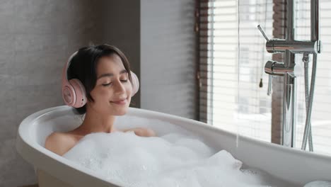 cheerful woman wearing headphones taking bath relaxing in bathroom indoors