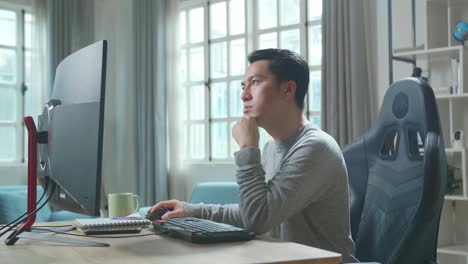 asian man in long sleeved t-shirt thinking then typing on desktop computer while working at home.