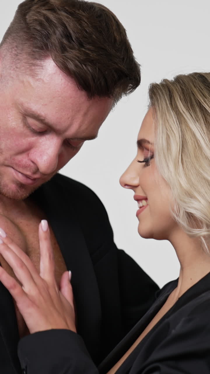 Two people in love wearing black jackets. Blonde long-haired woman stroking male chest and couple kissing. White backdrop. Vertical video